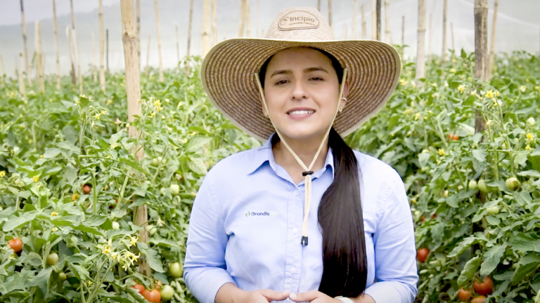 Mujer sonriente con sombrero en un campo de cultivo, con plantas verdes al fondo.
