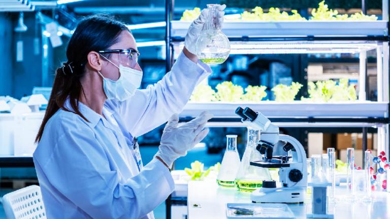 Científica con mascarilla analiza muestras en laboratorio con estantes iluminados de fondo.
